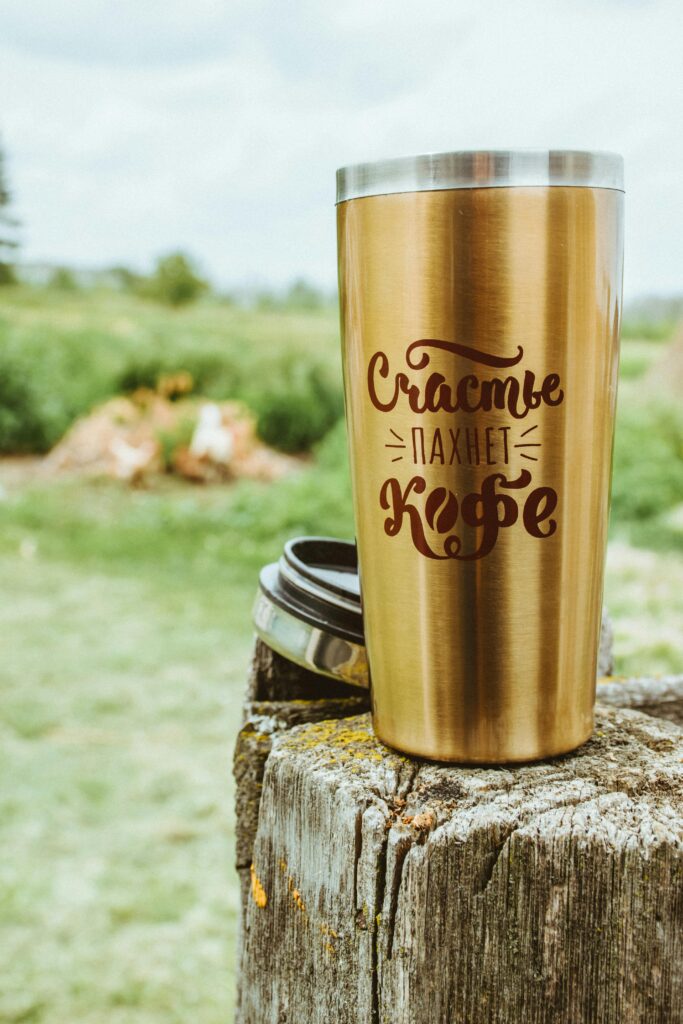 A gold travel tumbler with coffee-themed text on rustic wood outdoors.